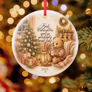 Könnte beinhalten: Ein runder, weißer Keramik-Weihnachtsanhänger mit rotem Band, bedruckt mit einem Teddybären mit Tasse, einem geschmückten Weihnachtsbaum, Geschenken und dem Text Frohe Weihnachten und ein glückliches neues Jahr 2026.