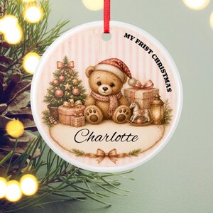 Könnte beinhalten: Ein rundes, weißes Keramikornament mit rotem Band, das einen braunen Teddybären mit Weihnachtsmütze und Schal, einen Weihnachtsbaum, Geschenke, ein Schaukelpferd, eine Laterne sowie den Text MY FRIST CHRISTMAS und Charlotte zeigt.