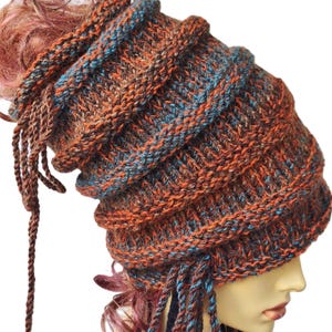 Peut inclure: Un bonnet tricoté à la main, multicolore, dans les tons orange, marron et bleu sarcelle. Le bonnet présente un design superposé unique et de longues tresses tressées. Un accessoire de mode original.