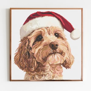 May include: A framed cross-stitch portrait of a dog wearing a Santa hat. The dog has light brown fur, a brown nose, and dark eyes. The Santa hat is red with a white brim and a white pom-pom.