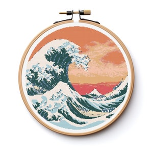 May include: A cross-stitch embroidery hoop featuring a rendition of The Great Wave off Kanagawa, a well-known Japanese woodblock print by Hokusai. The wave is depicted in shades of blue, green, and white, with a red and orange sunset in the background.