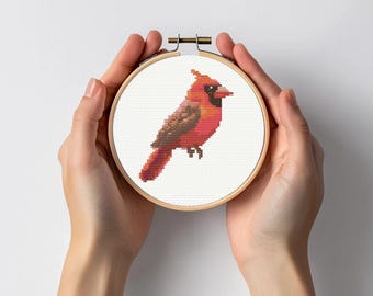 May include: A cross-stitch embroidery of a red cardinal bird on white fabric, set in a wooden hoop. The bird has brown and red details. The hoop is held by hands against a white background.