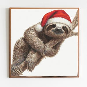 May include: A framed cross-stitch of a sloth wearing a red and white Santa hat, perched on a brown branch. The sloth has brown fur and is smiling. The artwork is displayed on a white background.