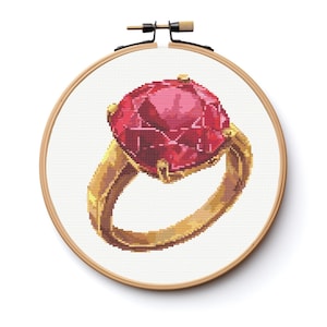 May include: Cross-stitch design of a gold ring with a large, faceted red gemstone. The ring is set against a white background within a wooden embroidery hoop, showcasing the detailed needlework and vibrant colors.