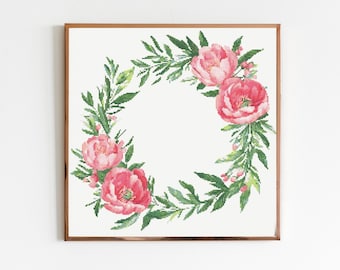 May include: A framed cross-stitch floral wreath featuring vibrant pink peonies and green leaves. The wreath is arranged in a circular pattern, with the flowers and foliage surrounding a white centre. The frame is a warm brown colour.