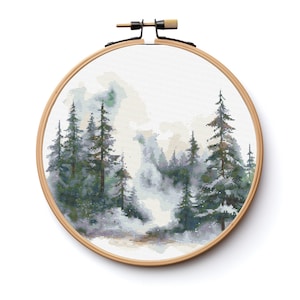 May include: A cross-stitch embroidery hoop featuring a winter forest scene. The design depicts a group of pine trees in shades of green and blue, against a white background with a hazy, foggy effect.