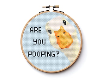 May include: A circular wooden embroidery hoop containing a cross-stitch pattern of a white goose with an orange beak on a light blue background. The black text on the left side of the fabric reads, ARE YOU POOPING? The design is centered.