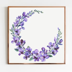 Puede incluir: Un diseño floral en punto de cruz enmarcado con una corona en forma de media luna de flores moradas y hojas verdes. La obra de arte está sobre un fondo blanco y enmarcada en un marco de madera marrón.