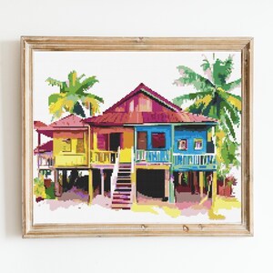 May include: A framed, colorful cross-stitch of a tropical house. The house is elevated on stilts and features a red roof, yellow and blue walls, and a staircase. Palm trees frame the house, and the artwork is in a wooden frame.