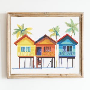 May include: A framed cross-stitch artwork depicts three colorful beach huts on stilts, each with a different color scheme: yellow, orange, and blue. Palm trees are visible above the huts. The artwork is framed in a light brown wooden frame.