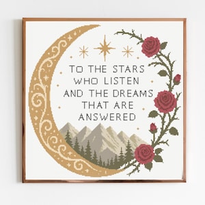 May include: A square cross-stitch pattern featuring a gold crescent moon, a mountain range with pine trees, and red roses on a vine. The text reads: TO THE STARS WHO LISTEN AND THE DREAMS THAT ARE ANSWERED. The design includes gold star accents.