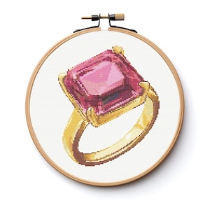 May include: Cross-stitch of a gold ring with a large, square, pink gemstone. The ring is set against a white background within a wooden embroidery hoop. The design features detailed stitching.