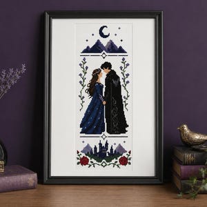 May include: A framed cross-stitch pattern featuring a couple standing face-to-face in a dark blue gown and black cloak. The design includes a crescent moon, mountain peaks, a castle silhouette, and red roses, all rendered in a pixelated style.
