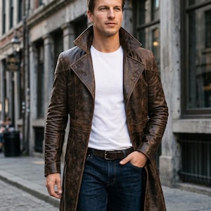 Peut inclure: Un long trench-coat en cuir marron vieilli avec un large col et des revers, porté sur un t-shirt blanc à col rond et un jean bleu foncé avec une ceinture en cuir marron, présentant une coupe structurée et des coutures apparentes.
