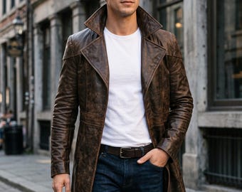 Puede incluir: Una gabardina larga de cuero marrón desgastado con cuello ancho y solapas, usada sobre una camiseta blanca de cuello redondo y pantalones vaqueros azul oscuro con un cinturón de cuero marrón, con ajuste estructurado y costuras visibles.
