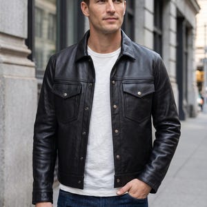 Könnte beinhalten: Eine schwarze Lederjacke mit klassischem Kragen, langen Ärmeln und zwei Brusttaschen mit Knopfverschluss. Die Jacke wird über einem schlichten weißen T-Shirt und einer dunkelblauen Jeans getragen und zeigt eine strukturierte Passform.