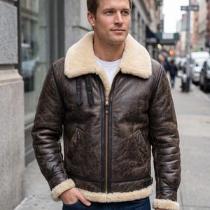 May include: A brown distressed leather jacket featuring a cream-colored shearling collar and lining. The jacket includes a central metal zipper, black leather trim details, and adjustable buckle straps on the chest and waist area.