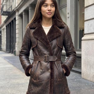 Peut inclure: Un trench-coat en cuir marron foncé doté d'une doublure en peau lainée épaisse et moelleuse, d'un large col cranté et d'une ceinture en cuir assortie avec une boucle rectangulaire. Le manteau présente un boutonnage croisé et des manches longues.