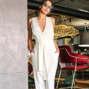May include: A woman wearing a white sleeveless jumpsuit with a wide belt and a deep V-neckline. She is wearing black sunglasses and has her hands in her pockets.