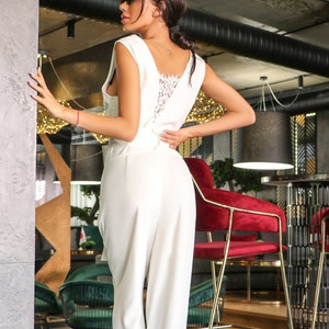 May include: A white sleeveless jumpsuit with a lace back detail. The jumpsuit is being worn by a person with dark hair and sunglasses. The person is standing in a restaurant setting.