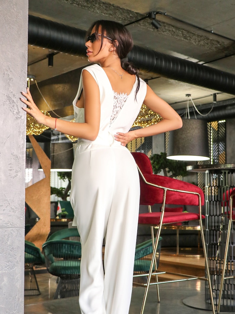 May include: A white sleeveless jumpsuit with a lace back detail. The jumpsuit is being worn by a person with dark hair and sunglasses. The person is standing in a restaurant setting.