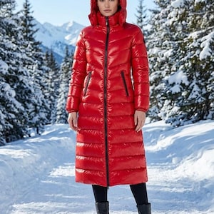 May include: A long, bright red puffer coat featuring a high collar, an attached hood, and a full-length black front zipper. The garment has horizontal quilted stitching, long sleeves with elasticized cuffs, and two zippered side pockets.