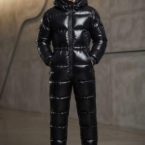 Puede incluir: Un mono de una pieza negro, acolchado y de alto brillo, con cremallera frontal completa, capucha, mangas largas con puños acanalados y bolsillos laterales. La prenda tiene un acabado brillante tipo plumas con costuras horizontales.