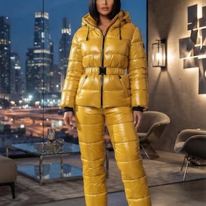 May include: A mustard yellow puffer jacket and matching puffer pants set with a glossy finish. The jacket features a hood, a black front zipper, and a black buckled belt at the waist. The pants have a high-waisted fit and elasticized cuffs.