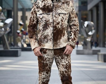 Op de afbeelding: Een bijpassende tweedelige set bestaande uit een jas met lange mouwen en een broek, beide gemaakt van bruin en wit koeienhuidleer met een natuurlijk vlekkenpatroon. De jas heeft een ritssluiting aan de voorzijde en twee borstzakken.