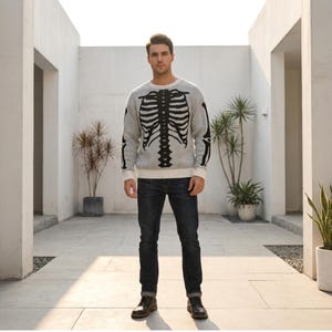 May include: A light gray long-sleeve crewneck sweater featuring a prominent black graphic print of a human rib cage on the front and arm bones on the sleeves, paired with dark blue denim jeans and dark brown leather boots.