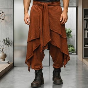 May include: A pair of rust-colored layered trousers featuring a tie-waist detail and a draped, asymmetrical hemline. The pants are paired with a dark gray t-shirt and dark brown leather lace-up boots, creating a layered look.