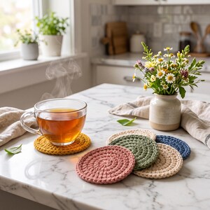 May include: A clear glass mug filled with amber tea sits on a round mustard yellow crocheted coaster. Nearby, a stack of four additional round crocheted coasters in dusty rose, sage green, cream, and navy blue are arranged on a white surface.