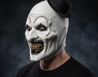 Könnte beinhalten: Eine Vollkopf-Clownmaske mit glatter weißer Oberfläche, einem kleinen schwarzen Melonen-Hut auf der Oberseite, geschwungenen schwarzen Augenbrauen, einer spitzen Nase und einem breiten Mund mit dunklen Lippen, die verfärbte, gelbe Zähne zeigen.