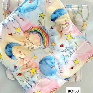 May include: A colorful fabric pot holder with a watercolor design of a rainbow, stars, moons, and hearts. The text "the world to me INE ou rand ways keep looking up there may RAO may the stars SHINE upon you forever and the world moon and back many the stars SHINE upon you forever and" is written on the fabric. The pot holder has a white background with a light blue, pink, and yellow watercolor design.