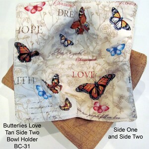 May include: A tan fabric bowl holder with a white fabric insert featuring a butterfly pattern and the words "HOPE", "DREAM", "LOVE", and "WITH". The bowl holder is labeled "Butterlies Love Tan Side Two Bowl Holder BC-31".