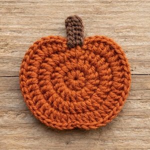 Può includere: Un sottobicchiere all'uncinetto piatto e circolare a forma di zucca, caratterizzato da un corpo in filato arancione brillante con un motivo a punti a spirale testurizzato e un piccolo stelo rettangolare marrone scuro in cima.