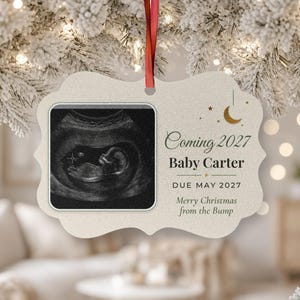 May include: A cream-colored, scalloped-edge ornament featuring a black and white ultrasound image on the left. The text reads Coming 2027, Baby Carter, DUE MAY 2027, and Merry Christmas from the Bump, with a small gold crescent moon icon.