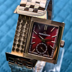 Op de afbeelding: Een goudkleurig rechthoekig polshorloge met een dieprode sunburst wijzerplaat, gouden indexen en wijzers. De wijzerplaat bevat de tekst Jaeger-LeCoultre en Swiss Made. Het horloge heeft een metalen schakelarmband met een textuur.