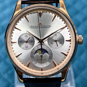 Può includere: Un orologio automatico Jaeger-LeCoultre con cassa in oro rosa, quadrante argentato a raggiera e cinturino in pelle nera. Il quadrante presenta tre sotto-quadranti, un indicatore delle fasi lunari e indici dorati. Il testo riporta Jaeger-LeCoultre e Swiss Made.