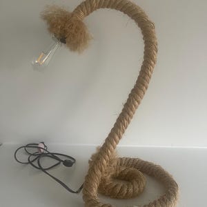 May include: A unique table lamp crafted from thick, coiled rope. The lamp features a curved design with a lightbulb at the top and a power cord. The rope is a natural beige color, creating a rustic and textured appearance.