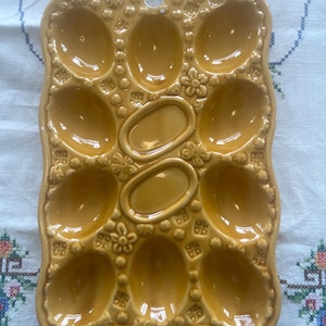 Pode incluir: Uma bandeja de ovos de cerâmica amarela mostarda vintage com um design floral decorativo e em relevo. A bandeja tem 12 compartimentos ovais para ovos, mais dois compartimentos ovais menores. Há um pequeno orifício na parte superior para pendurar.