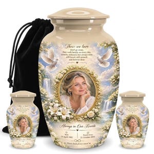 May include: A large and two small cream-colored cremation urns featuring a central oval portrait frame surrounded by white flowers, two flying doves, and a waterfall. The text reads Those we love don't go away, Always in Our Hearts.
