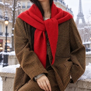 May include: A bright red knit scarf draped over the shoulders of a brown and black plaid blazer. The blazer features a buttoned front and long sleeves, worn over a light-colored shirt. The scarf is tied in a knot at the center of the chest.