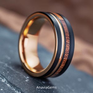 May include: A ring featuring a black brushed metal exterior, a central inlay of brown wood grain, and a polished rose gold interior. The text AnaviaGems is printed at the bottom of the image. The band has a smooth, rounded profile.