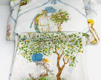 Può includere: Un set di lenzuola in tessuto bianco con illustrazioni di un personaggio con cuffia blu. Una sezione mostra il personaggio che cura un cespuglio di rose sotto un nastro blu, l'altra su un'altalena sotto un albero di limoni.