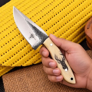 May include: A fixed-blade knife with a silver steel blade featuring a dark, textured spine and a laser-etched deer skull logo. The handle is made of smooth, cream-colored material with a black deer head graphic and two silver rivets.