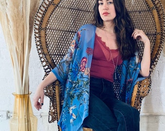 Può includere: Una donna con un kimono floreale blu, un top bordeaux e jeans neri, seduta su una sedia a pavone in vimini. Alte erbe secche in vasi color ambra su entrambi i lati. Il pavimento è in legno chiaro.