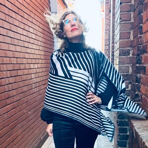Op de afbeelding: Een vrouw draagt een zwart-wit gestreepte poncho met een zwarte turtleneck en een zwarte flared broek. Ze staat in een bakstenen steegje en kijkt omhoog.