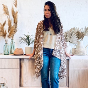 May include: A woman wearing a brown and white patterned kimono, blue jeans, and brown boots. She is standing in front of a wooden dresser with a rug on the floor.