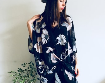 May include: A woman's outfit featuring a tan fedora hat and a black floral kimono-style top. The top has a white and pink floral pattern. The outfit is paired with black pants. A small potted plant is in the background.
